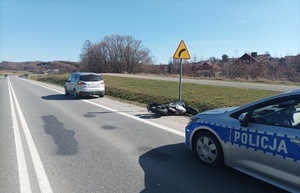 Kolizja motocykla z osobowym fordem. Na miejscu policja drogowa, oznakowany radiowóz z napisem Policja.