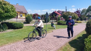 Uczestnik turnieju BRD jadący rowerem przez miasteczko ruchu drogowego wraz z policjantem sprawdzającym jego umiejętności i znajomość przepisów.