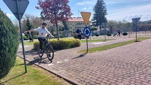 Uczestnik turnieju BRD jadący rowerem przez miasteczko ruchu drogowego wraz z policjantem sprawdzającym jego umiejętności i znajomość przepisów.