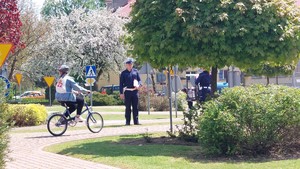 Uczestnik turnieju BRD jadący rowerem przez miasteczko ruchu drogowego wraz z policjantem sprawdzającym jego umiejętności i znajomość przepisów.