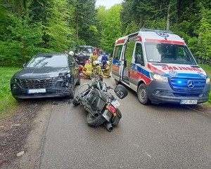 Na pierwszym planie widać pojazdy uczestniczące w zdarzeniu drogowym oraz karetkę pogotowia, w tle strażacy oraz ratownicy medyczni udzielający pierwszej pomocy poszkodowanemu, a za nimi policjant i radiowóz.