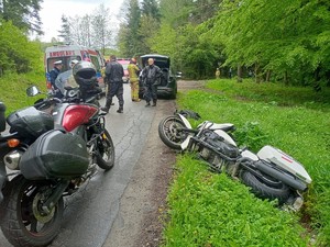 Na pierwszym planie widać stojący motocykl oraz drugi motocykl leżący na boku poza drogą. W tle służby ratownicze i uczestnicy zdarzenia.