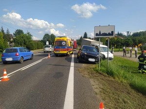 Po lewej stronie zdjęcia widać pojazdy omijające miejsce zdarzenia drogowego. W środkowej części zdjęcia widoczna karetka pogotowia a po jej prawej stronie za znakiem drogowym oznaczający obszar zabudowany widać uczestniczące w zdarzeniu pojazdy. Najpierw widać pojazd marki KIA a za nim samochód marki Iveco. Po prawej stronie zdjęcia osoby obserwujące miejsce zdarzenia i grupę umundurowanych strażaków biorących udział w obsłudze zdarzenia.