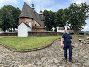 Na zdjęciu na pierwszym planie widać umundurowanego policjanta, który stoi przed kościołem w miejscowości Blizne, będący jednocześnie zabytkiem wpisanym na światową listę UNESCO. W tle zdjęcia widoczna zabudowa sakralna z otaczającym kościół murem oraz sam budynek kościelny.