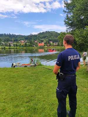 Na zdjęciu widać stojącego tyłem umundurowanego policjanta przed zbiornikiem wodnym w Krzemiennej przy ośrodku wypoczynkowym &quot;U Schabińskiej - Krzemienna nad Sanem&quot;. Na zbiorniku wodnym widać jak strażacy z Komendy Powiatowej Państwowej Straży Pożarnej w Brzozowie poruszają się na motorówce, co jest częścią pokazu ratownictwa wodnego dla biorących udział w spotkaniu.