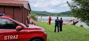 Na zdjęciu po lewej stornie oznakowany wóz bojowy straży pożarnej. Dalej na środku stoi tyłem dwóch policjantów umundurowanych, którzy brali udział w spotkaniu profilaktycznym. Następnie po lewej stronie widać część domku letniskowego i weranda, na której znajdują się dzieci i opiekunowie słuchający prelekcji na temat Bezpieczeństwa nad Wodą. W dalszym tel zdjęcia w środkowej części zdjęcia stoi ubrany w specjalistyczny mundur strażak KP PSP w Brzozowie, który w tym momencie prowadzi spotkanie. W samym tle zdjęcia widoczny pomost idący w głąb zbiornika i sam zbiornik wodny, za tym awenem widoczne tereny leśne na terenie górzystym.