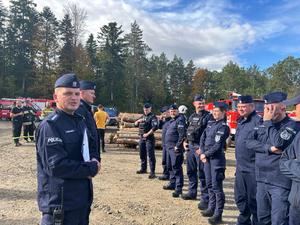 Zdjęcie przedstawia przeprowadzaną na miejscu ćwiczenia odprawę przeprowadzaną dla policjantów przez dowódców. W tle widoczni są druhowie z OSP, strażackie wozy bojowe, składowane drewno oraz teren leśny.