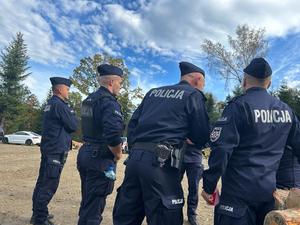 Na zdjęciu widoczni odwróceni plecami umundurowani funkcjonariusze policji. W tle po lewej stronie widać samochód osobowy koloru białego, a w tle kadru widać drzewa oraz chmury na niebie.