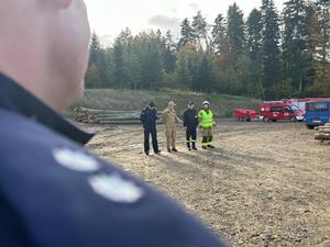 Zdjęcie w pierwszym tle kadru ukazuje ramię policjanta z oznaczeniem stopnia policyjnego na pagonie podkomisarza, jest to rozmazany obraz. W tle widoczni dowódcy szkolenia przeprowadzający szkolenie. Od lewej: młodszy inspektor Marek Śliwiński Zastępca Komendanta Powiatowego Policji w Brzozowie, brygadier Bogdan Biedka Zastępca Komendanta Powiatowego Państwowej Straży Pożarnej w Brzozowie, druh Marek Owsiany Prezes Zarządu Gminnego Zastępów Ochotniczych Straży Pożarnych, druh Adam Toczek Komendant Gminny Ochrony Przeciwpożarowej. Dalej na końcu kadru wdidać ustawione strażackie wozy bojowe, ułożone drewno oraz teren leśny.