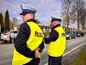 Na zdjęciu w pierwszym kadrze widoczni są umundurowani policjanci ruchu drogowego z kamizelkami odblaskowymi z napisem policja stojący plecami. Policjant po lewej stronie patrzy w lewym kierunku drogi kontrolując sytuację ruchu drogowego. W tle zdjęcia widać cmentarz w miejscowości Przysietnica a przed nim zaparkowane wzdłuż samochody osobowe.