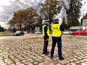 Na pierwszym planie widoczni są dwaj umundurowani policjanci ruchu drogowego z ubranymi kamizelkami odblaskowymi, którzy obserwują sytuację drogową w rejonie cmentarza komunalnego w Brzozowie stojąc na rondzie. W tle widoczne samochody osobowe poruszające się po drodze. Dalej w kadrze widać ogrodzenie cmentarza a na końcu kadru po lewej stronie widoczny szyld stacji paliw.