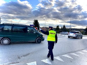 Zdjęcie przedstawia na pierwszym tle umundurowanego policjanta ruchu drogowego z założoną kamizelką odblaskową z napisem policja, który reguluje ręcznie ruch odbywający się na rondzie w Brzozowie. Przed policjantem i po jego prawej stronie widoczne są pojazdy osobowe. Na środku zdjęcia za pojazdem widać oznakowanie drogowe, latarnie uliczne, a tło stanowi zachmurzone niebo.