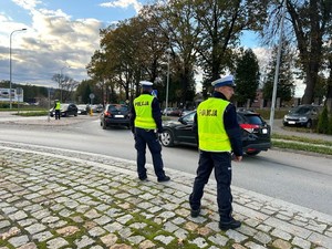 Na pierwszym planie widoczni są dwaj umundurowani policjanci ruchu drogowego z ubranymi kamizelkami odblaskowymi, którzy obserwują sytuację drogową w rejonie cmentarza komunalnego w Brzozowie stojąc na rondzie. W tle widoczne samochody osobowe poruszające się po drodze. Dalej w kadrze po lewej stronie widać kolejnego umundurowanego policjanta ruchu drogowego z kamizelką odblaskową kierującego ruchem drogowym. W tle widać ogrodzenie cmentarza a tło zdjęcia stanowią zadrzewienia i zachmurzone niebo.