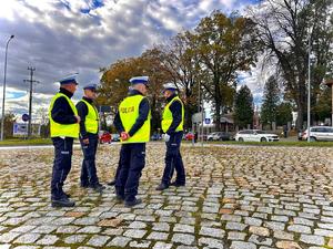 Na pierwszym planie widoczni są czterej umundurowani policjanci ruchu drogowego z ubranymi kamizelkami odblaskowymi, którzy obserwują sytuację drogową w rejonie cmentarza komunalnego w Brzozowie stojąc na rondzie. W tle widoczne samochody osobowe poruszające się po drodze. Dalej w kadrze widać ogrodzenie cmentarza. Na zdjęciu w tle widoczne zadrzewienia oraz zachmurzone niebo.