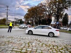 Na zdjęciu od prawej na pierwszym planie widoczny samochód osobowy koloru białego zatrzymywany na drodze przez stojącego po lewej stronie umundurowanego policjanta ruchu drogowego ubranego w kamizelkę odblaskową z napisem policja, który kieruje ruchem drogowym. Policjant ten wykonuje gest wskazujący miejsce, w którym samochód ma się zatrzymać. Dalej w kadrze widoczne samochody osobowe jadące drogą. Po prawej stronie w tle widoczni są piesi czekający na możliwość przejścia przez drogę a od nich po lewej stronie widać głowę umundurowanego policjanta ruchu drogowego. Dalej w tle widoczne ogrodzenie cmentarza komunalnego w Brzozowie a tło zdjęcia stanowią drzewa oraz zachmurzone niebo.