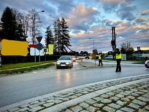 Od prawej strony widoczny umundurowany policjant ruchu drogowego ubranego w kamizelkę odblaskową, który reguluje ręcznie ruch odbywający się na rondzie w Brzozowie w rejonie cmentarza komunalnego. Drugi policjant stoi w rejonie przejścia dla pieszych. Następnie po lewej stronie widać samochody osobowe stojące jadące z kierunku miejscowości Przysietnica, a które właśnie są wpuszczane na skrzyżowanie przez policjanta. Po lewej stronie w tle widoczne zamazane banery reklamowe. Tło stanowią po lewej stronie drzewa a na całym zdjęciu zachmurzone niebo.