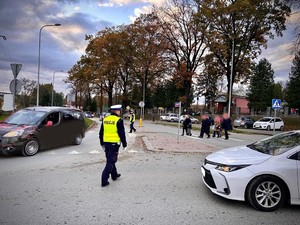 Od prawej strony widać przód samochodu osobowego koloru białego, dalej po lewej stronie na środku zdjęcia widoczny umundurowany policjant ruchu drogowego z ubraną kamizelką odblaskową z napisem policja, który stoi na drodze i reguluje ruch drogowy. po jego lewej stronie widać kolejny samochód, który wjeżdża na skrzyżowanie o ruchu okrężnym. W tle wioczny jest drugi umundurowany policjant ruchu drogowego z ubraną kamizelką odblaskową, który również kieruje ruchem drogowym i widoczni są po prawej stornie przechodzący przez przejście dla pieszych piesi. W tle widoczne po prawej stronie zaparkowane przed cmentarzem komunalnym w Brzozowie samochody osobowe, a dalej widać teren cmentarza wraz z kaplicą cmentarną. W tle widać od prawej strony drzewa a dalej w lewo widać zachmurzone niebo.