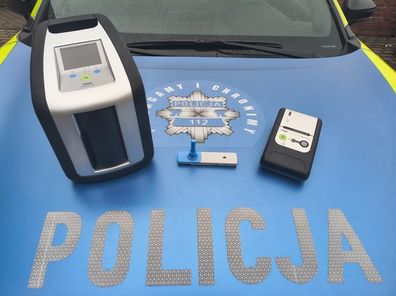 Na zdjęciu widoczne urządzenie Drug Test - pozwalające sprawdzić w próbce śliny substancji narkotycznych. Zdjęcie wykonane na pokrywie pojazdu służbowego, widoczny napis POLICJA.